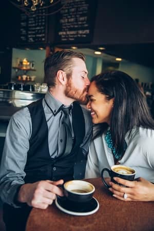 a man in a suit kissing his girlfriend’s forehead in a cafe