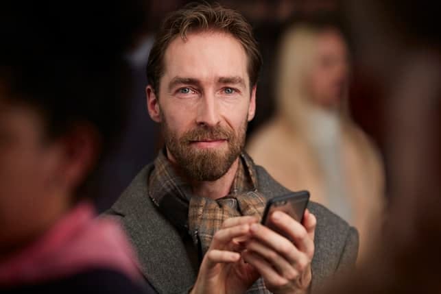 a mature man with sad eyes is looking for someone in the crowd while holding a smartphone in his hand