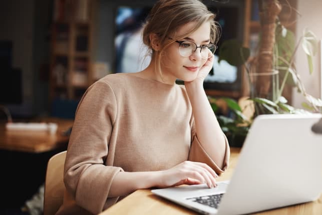 a young girl in glasses is using her laptop while smiling