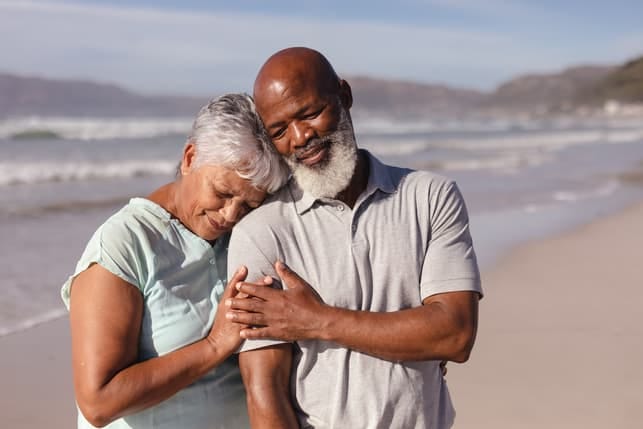 a black senior couple is cuddling while having a date on a beach
