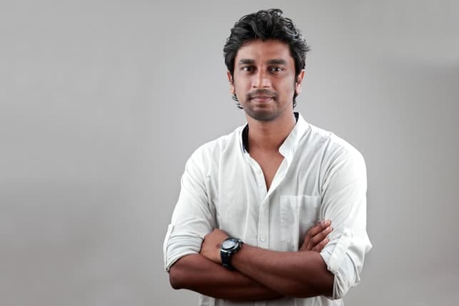 a handsome indian man wearing a white shirt and posing with crossed hands
