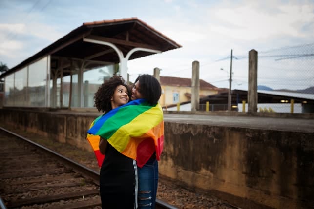 two lesbians hugging and posing with a rainbow flag on a railway station